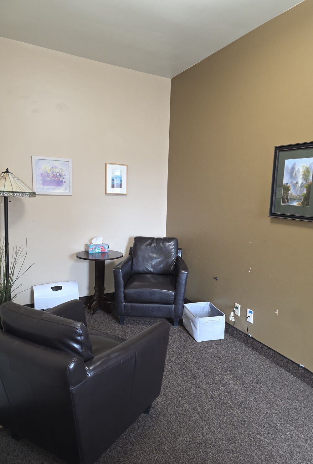View upon entering therapy space. Image shows 2 chairs, angled towards each other, a gray basket, artwork featuring plants, and a lamp. 
