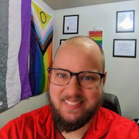 Gallery Photo of Daniel, a bald, white man sits in a corner near an intersex inclusive progress pride flag and asexual flag. The other wall has degrees and licenses.